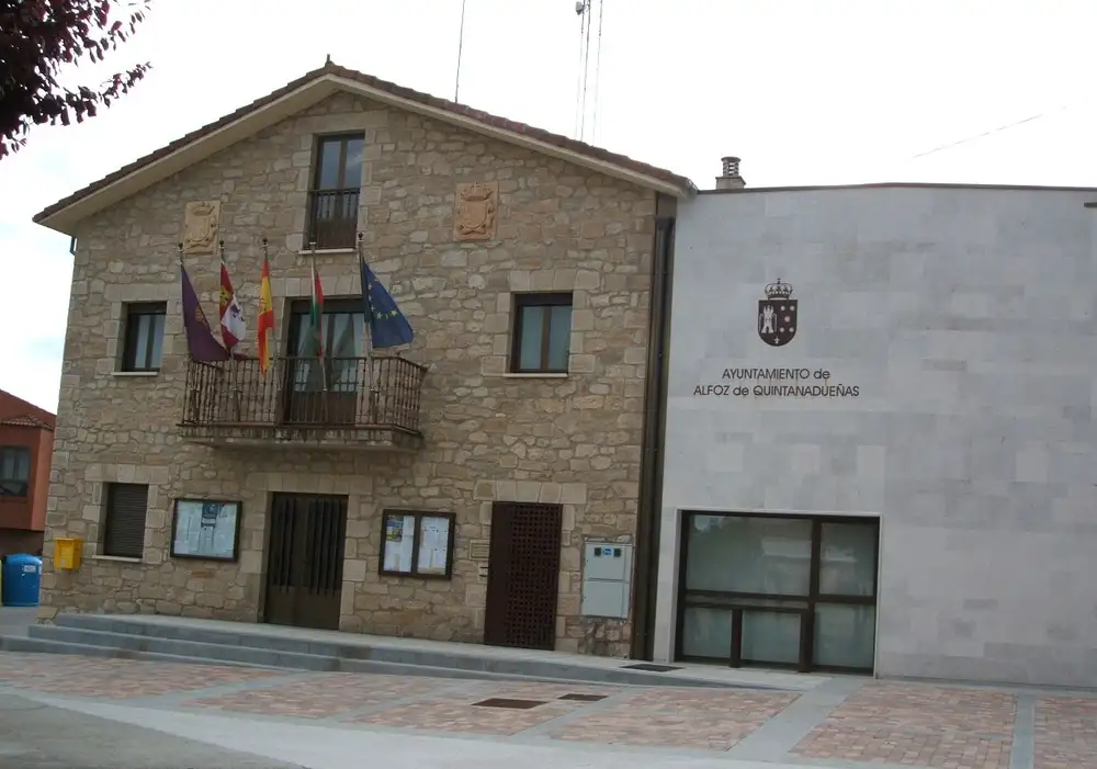 Casa consistorial de Alfoz de Quintanadueñas, un edificio de piedra de dos plantas con balcón y banderas, junto a un anexo moderno
