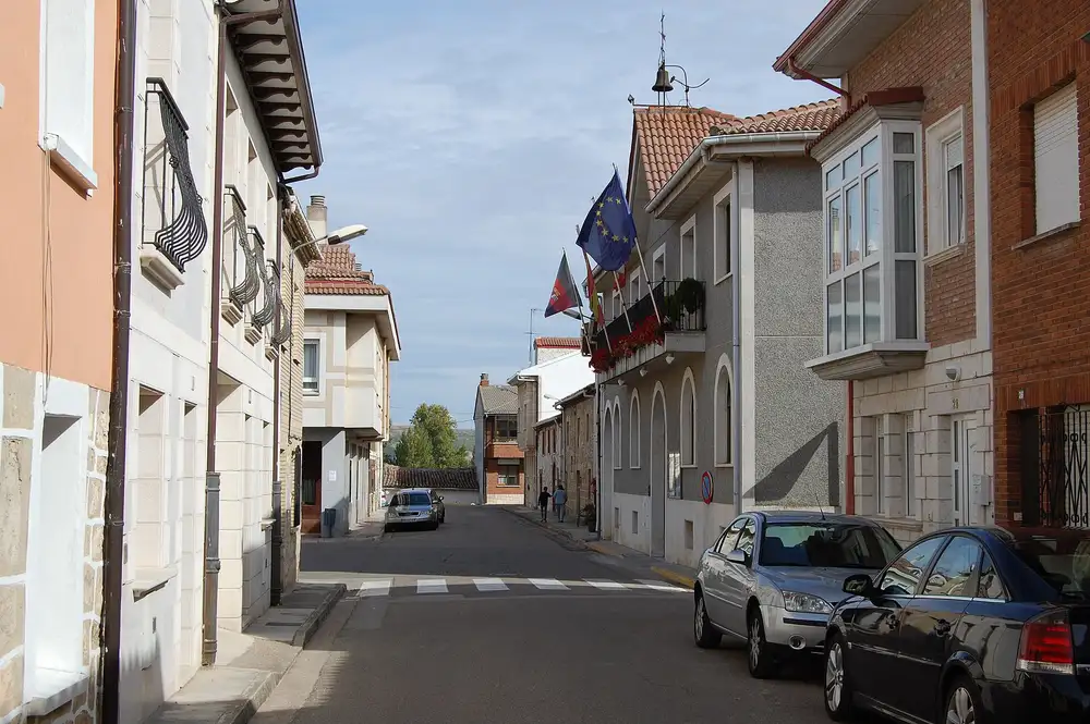 Calle de Villalbilla de Burgos con casas de dos y tres plantas, coches aparcados y banderas en la fachada del Ayuntamiento